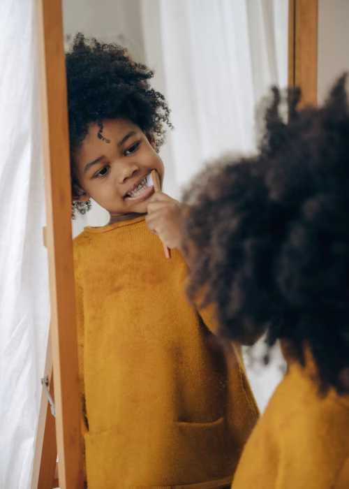 A young child smiling and checking her teeth in a mirror during her morning brushing routine.